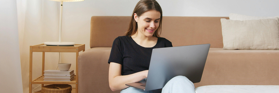 Mulher sentada no chão de sua sala usa o computador. Ela checa os resultados do sisu.