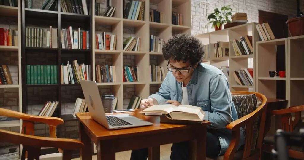 rapaz estudando com livros e computador em biblioteca